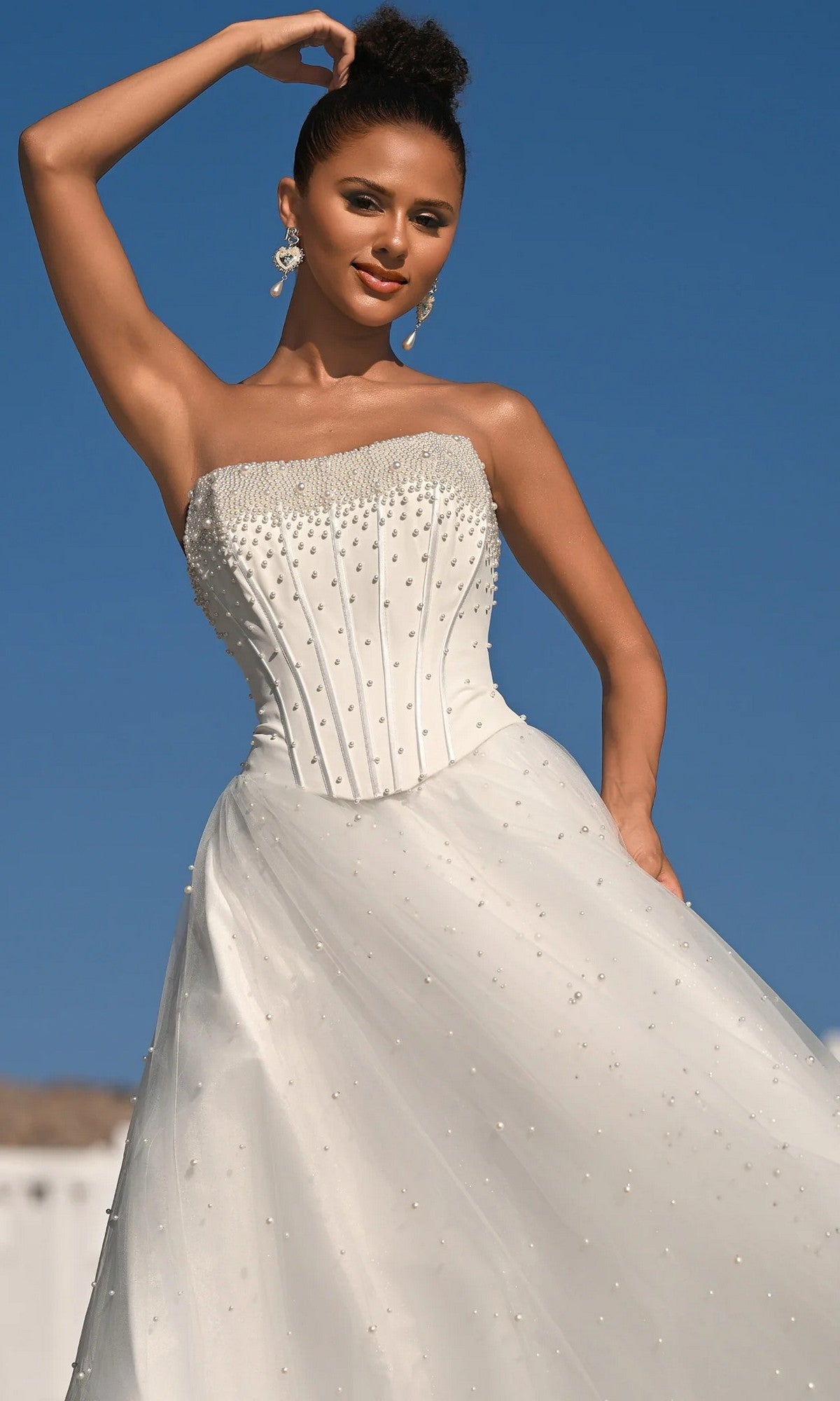 A woman wears the Portia and Scarlett PS26034 long formal dress, featuring a strapless white corset bodice with pearl embellishments, as she poses outdoors against a clear blue sky.